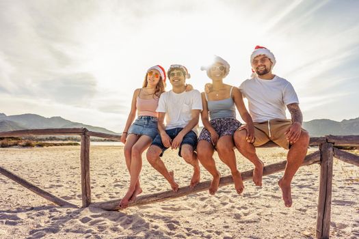 Suggestive photography with backlight sun effect of four young happy people enjoying outdoor life in exotic vacation resort sitting on wooden fence on a white sand beach at sunset