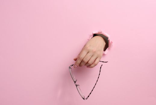 female hand holding glasses, part of body sticking out of torn hole in pink paper background