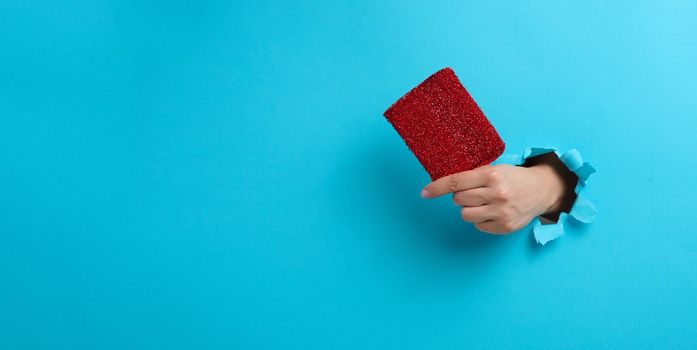 female hand holds a red kitchen sponge. Part of the body sticking out of a torn hole in a blue paper background