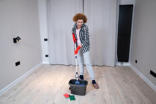Happy mature woman mops the floor and dances and sings in a new unfurnished apartment before moving to a new house. Housewife enjoying domestic chores, doing home cleanup creatively