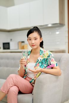 Happy woman taking vitamin pill holding glass sitting on a couch at home