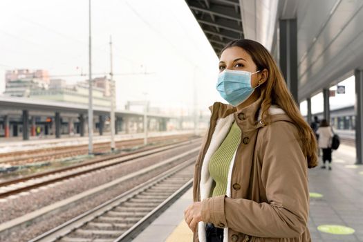 Beautiful woman in a medical mask waiting train on station. Portrait of a young woman with a surgical mask on her face against SARS-cov-2 outdoors.