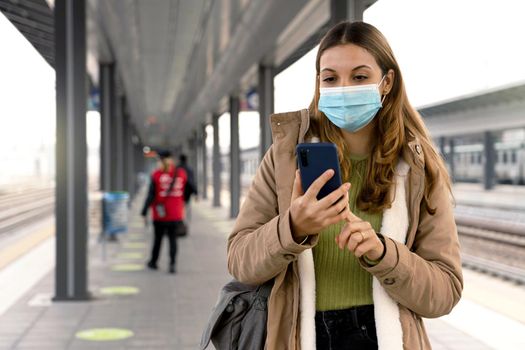 Beautiful young woman wearing medical mask ready to shows her sanitary passport on smartphone to get on the train at station outdoors
