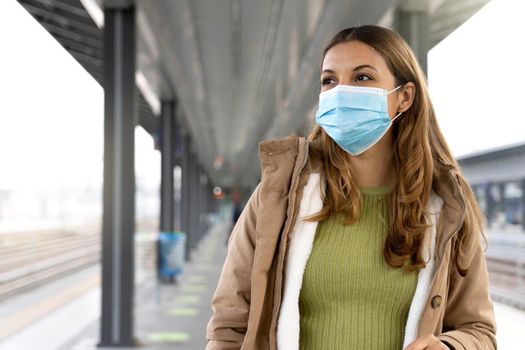 Commuter woman wearing surgical mask waiting train at station outdoors. Copy space.