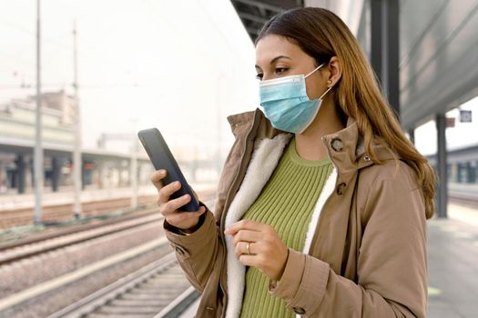 Portrait of beautiful woman with surgical mask using her smart phone waiting her train at station outside