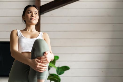 Portrait of asian girl doing workout at home, lifting legs and looking at distance, exercising in living room without fitness gear