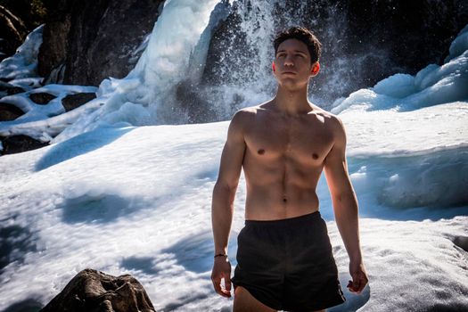 Shirtless athletic young man standing near snowy waterfall