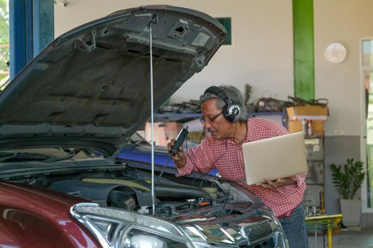 Asian automotive mechanic with red shirt use electronic tools and laptop to check the car in part of engine near hood in the garage.