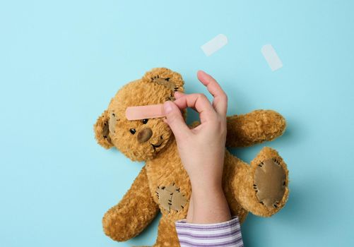 female hand holds a brown teddy bear and glues a medical adhesive plaster on a blue background, tram treatment