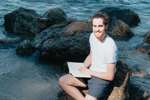 Young man smiling to camera while reading a book on the shore of the sea, copy space, confidence and study concepts, future and self help