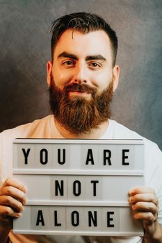 Young bearded hipster man holding a sign that says you are not alone, while smiling to camera, mental health and depression support concepts