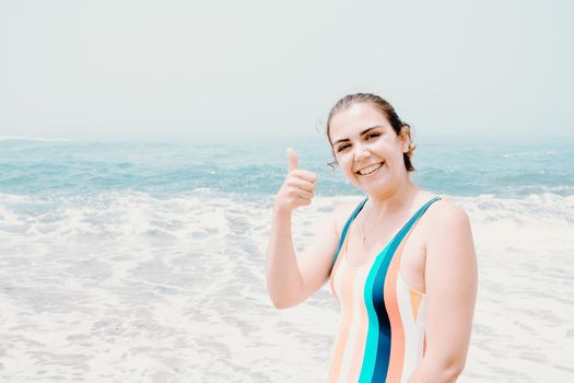 Young woman in a swimsuit salutes to camera during a sunny day at the beach with the ocean as background, liberty and holiday concept, copy space