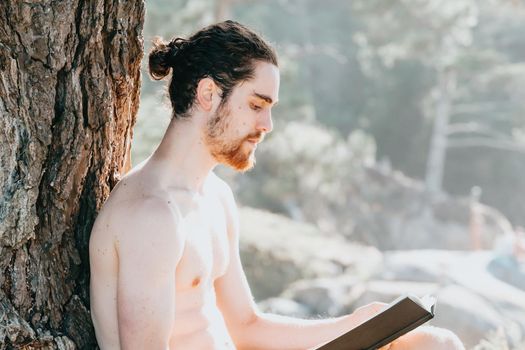 Close up of a man with a beard reading a book shirtless next to a tree during summertime