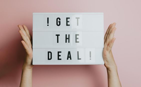 Two hands holding a sign that says get the deal over a pastel pink background, styling and design concept, copy space, buy online and e-commerce