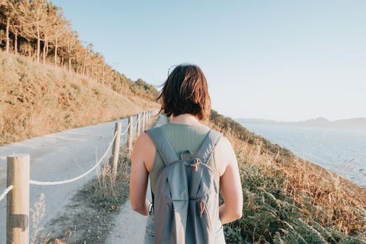 Back image of a young backpack traveler hiker during an stunning sunset coastline scenario, hipster traveler concept, beauty landscape. Copy space for text. Movement and liberty, nomad life.