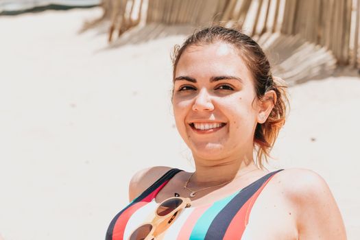 Young woman smiles to camera while wearing a colorful swimsuit at the beach, travel young holiday concept, copy space, social network, sunglasses user concept