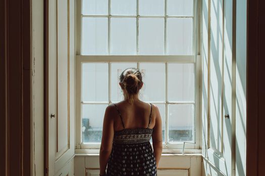 Young woman using a dress looking through a window, giving the back to the camera, stress and anxiety concepts, dark image, sadness