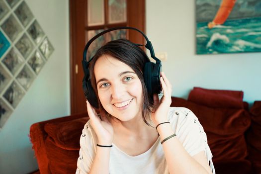 Portrait of a young playful hipster american woman in headphones smiling to camera in living room at home