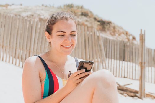 Young woman smiles while chatting on the phone wearing a colorful swimsuit at the beach taking a sun bath, travel young holiday concept, copy space, social network, sunglasses user concept