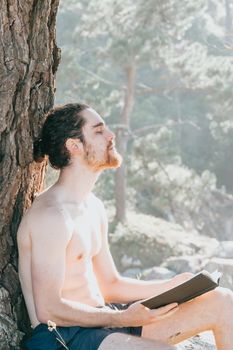 Close up of a man with a beard holding a book and relaxing shirtless next to a tree during summertime, vertical image
