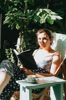 Woman holding a book looking serious to camera while reclined on a chair during a sunny day, copy space, relax and hobby concepts