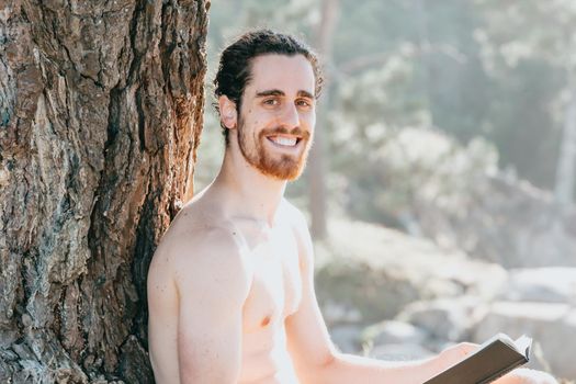 Close up of a man with a beard holding a book and smiling to camera shirtless next to a tree during summertime
