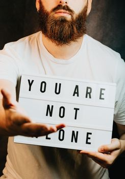 Young bearded hipster man holding a sign that says you are not alone, giving hands to the camera, help and self help concept, mental health