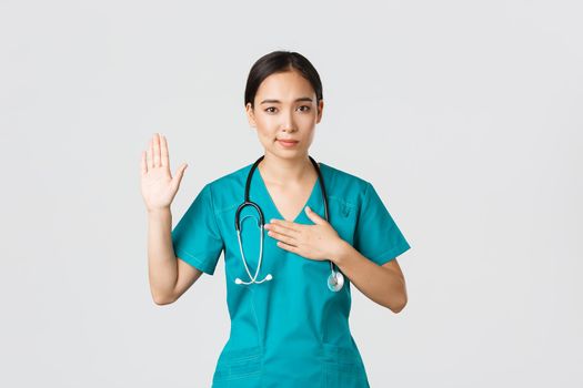 Covid-19, healthcare workers and preventing virus concept. Smiling honest asian female doctor, intern in scrubs giving pledge, raising one arm and hold hand on heart while promise, oath to patient
