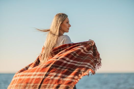 Attractive blonde Caucasian woman enjoying time on the beach at sunset, walking in a blanket and looking to the side, with the sunset sky and sea in the background. Beach vacation