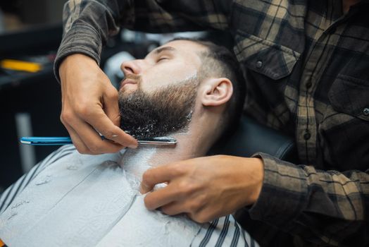 Young bearded man getting shaved by hairdresser at barbershop