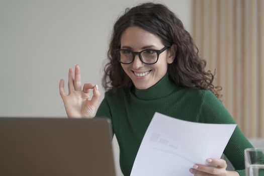 Smiling Italian female employee holding financial report and showing okay gesture during video call