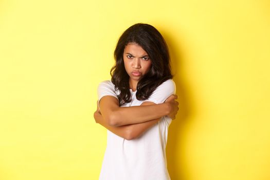 Image of moody cute african-american girl, hugging herself and sulking with offended expression, standing over yellow background