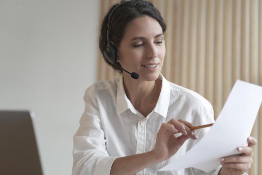 Positive Spanish businesswoman in headset sits at desk having video conference online with partners