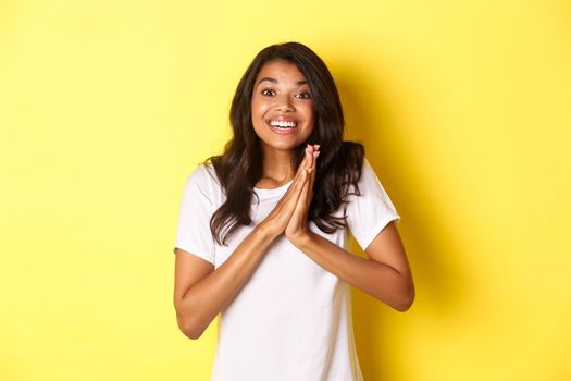 Image of happy and pleased african-american girl, clap hands and looking excited, thanking for something, standing over yellow background