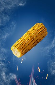 Grilled ear of corn hovering over flames in sparks and haze on blue background