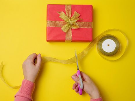 female hands are cutting a silk ribbon with scissors, next to gift boxes. Yellow background, top view