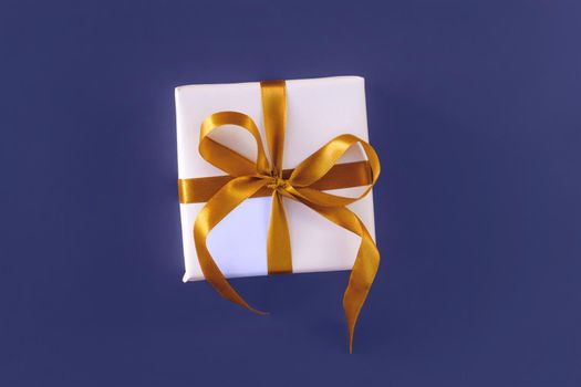 Gift box wrapped in white paper with a golden bow on festive violet blue background