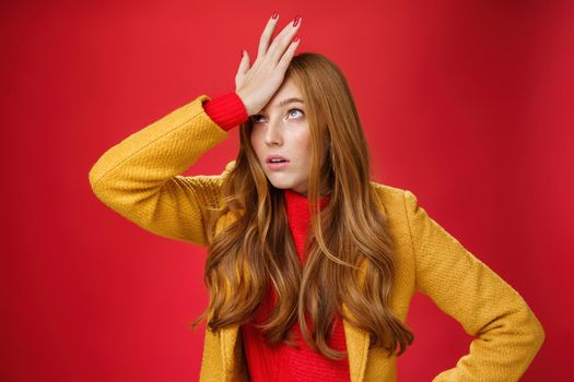 Redhead businesswoman making facepalm gesture with hand on forehead rolling eyes up from annoyance and irritation as being shocked with how dumb client is sighing bothered over red wall, tired