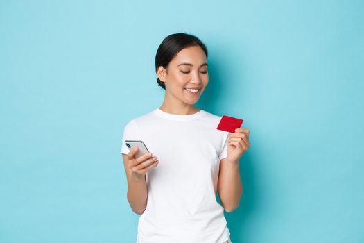E-commerce, shopping and lifestyle concept. Smiling attractive asian girl order online, looking at credit card while entering digits to mobile phone app, standing light-blue background
