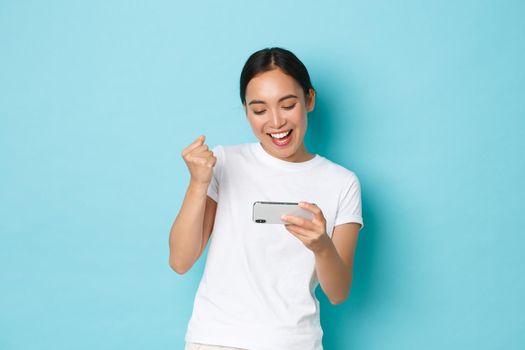Lifestyle, technology and people concept. Joyful pretty asian girl in white t-shirt having fun playing mobile arcade, winning in game, fist pump and looking satisfied at phone screen, blue background.