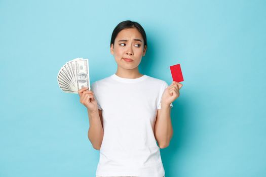 Shopping, money and finance concept. Indecisive and confused attractive asian girl cant decide whats better, credit card or cash, looking perplexed, standing thoughtful over blue background