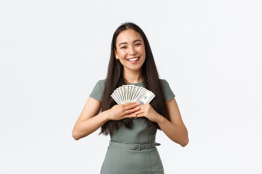 Gorgeous asian woman winning prize, holding money and smiling, thinking what to buy, earn cash for her startup, small business owner delighted after receiving first check, white background