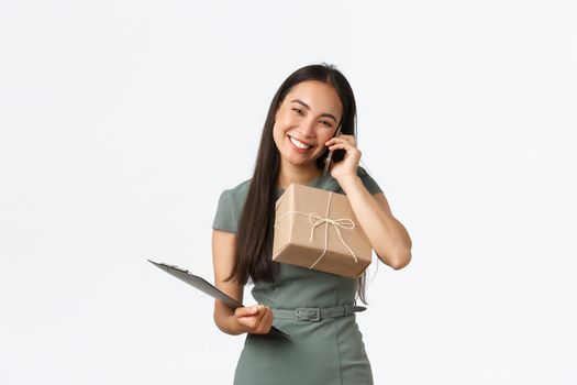 Friendly smiling and polite asian female shop owner, managing small business, carry clipboard and package box for shipping, answering clients call, talking with customer, checking info for delivery