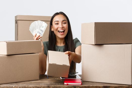 Small business owners, startup and e-commerce concept. Smiling asian woman showing money and giving box with order, selling online, earn cash for internet store, shipping handmade products