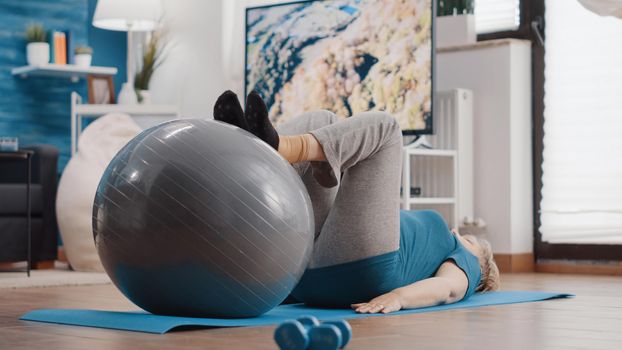 Old woman stretching legs muscles on fitness toning ball to exercise on yoga mat. Retired person training with sport equipment and doing physical activity for wellness at home. Senior adult