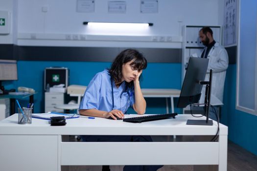 Sleepy nurse looking at computer screen for healthcare at office. Exhausted woman working late at night with monitor and technology for medical support. Tired assistant in cabinet