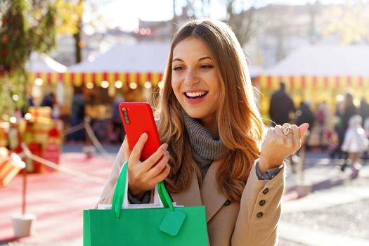 Attractive excited young woman watching smartphone and holding shopping bags walking in the Christmas Markets