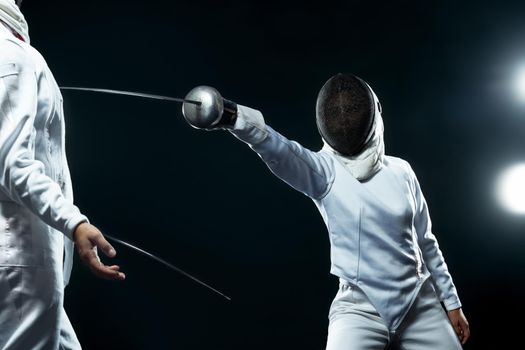 Two fencers on professional sports arena. Young fencer athlete wearing mask and white fencing costume. holding the sword on black background with lights.