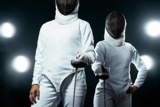 Young fencer athlete wearing mask and white fencing costume. holding the sword on black background with lights.