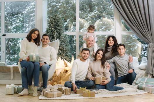 Portrait of noel large family gathering. One big stylish family. Grey-haired grandparents, granddaughter, grandson, brothers, sisters, grandchildren sitting on floor, getting gifts. Fir tree. Christmas mood